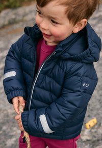Ungt barn i en marineblå dunjakke holder en stik udendørs på en klippeoverflade.
