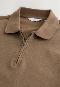 Tricou polo cu fermoar maro, cu o suprafață texturată, având un guler pliabil și un fermoar argintiu. Eticheta spune "NEXT."