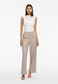 Λευκή αμάνικη μπλούζα με καπιτονέ σχέδιο, συνδυασμένη με ανοιχτό καφέ παντελόνι με ψηλή μέση και φαρδιά πόδια, και μπεζ γόβες με λουράκια.