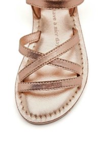 Sandalia metálica de oro rosa con tiras entrecruzadas, suela plana y detalles cosidos en los bordes. Textura suave y diseño minimalista.