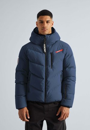 Marineblaue Steppjacke mit hohem Kragen und abnehmbarem Kapuzen, ausgestattet mit Reißverschlusstaschen und einem kontrastierenden roten Logo auf der linken Brust.