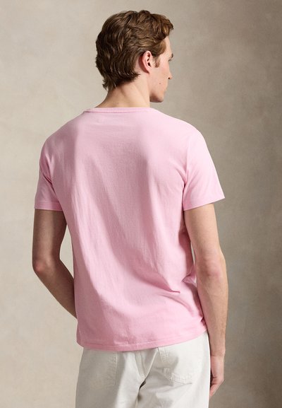 Mand vendt væk iført en ensfarvet lyserød T-shirt med korte ærmer og off-white bukser mod en neutral baggrund.