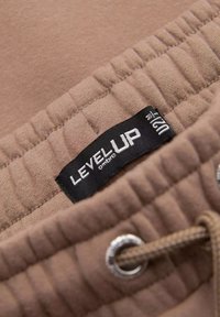 Beigefarbener Stoff mit einer weichen Textur, ausgestattet mit einem elastischen Bund, einer Metallöse und einem Etikett mit der Aufschrift "LEVEL UP" in Schwarz und Weiß.