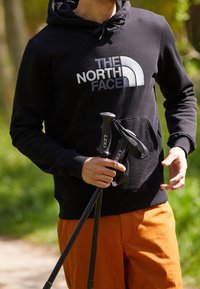 Černá mikina s kapucí a logem "THE NORTH FACE", obsahující klokaní kapsu. Osoba drží turistické hole Leki. Oranžové kalhoty, venkovní prostředí.