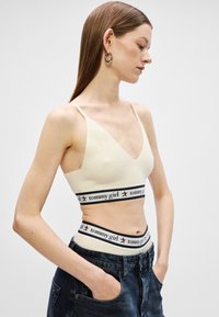Krémová rebrovaná braletka s trojuholníkovými poháre, široký elastický pás s logom "tommy girl" a akcentmi v čiernej a červenej farbe. Kombinovaná s džínsy so zvýšeným pásom.