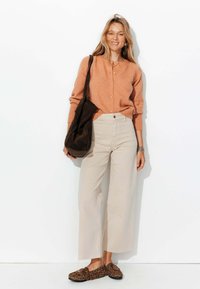 Gilet orange clair avec des boutons, pantalon large crème, sac épaule marron foncé et ballerines à motif animal. Textures lisses et coupe décontractée.