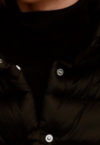 Zwarte pufferjas met glanzende knopen, voorzien van een doorgestikt ontwerp en een nauwsluitend hoogsluitend gebreide trui eronder. Soepele textuur.