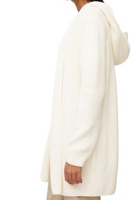 Boční pohled na osobu oblečenou v dlouhém, krémově bílém pleteném kardiganu s kapucí a béžových kalhotách.