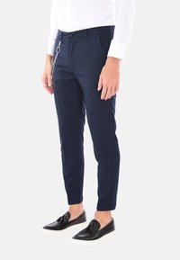 Pantaloni slim-fit blu navy realizzati in tessuto leggero, con dettaglio a catena in metallo, tasche laterali e orli arrotolati. Abbinati a scarpe nere slip-on.