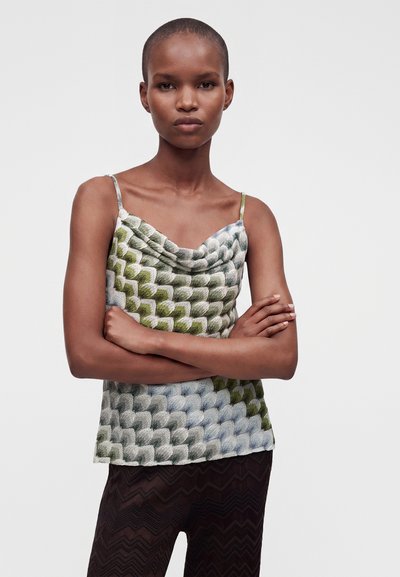 Femme aux cheveux courts croisant les bras, vêtue d'un haut sans manches texturé vert et gris à motifs et d'un pantalon sombre à motif zigzag.