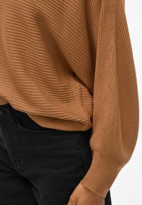 Pull à côtes brun avec une coupe ample, dotée de manches longues et d'une finition texturée. Associé à un jean noir.