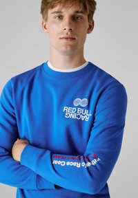 Νεαρός άνδρας με ανοιχτόχρωμα μαλλιά φορώντας μια μπλε φανέλα Red Bull Racing με τα χέρια σταυρωμένα μπροστά σε απλή γκρι φόντο.