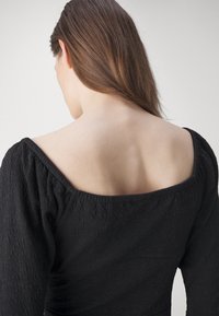 Black textured garment with puffed long sleeves and a wide neckline, showcasing the back and upper neckline. Smooth skin visible.