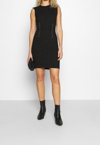 Robe noire ajustée sans manches, avec des coutures courbées et des clous décoratifs. Assortie à des bottines noires et un petit sac à main.