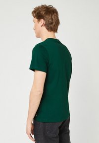 Jonge man met lichte huid en bruin haar, gekleed in een donkergroen T-shirt met korte mouwen en zwarte jeans, staand met zijn gezicht naar links tegen een witte achtergrond.