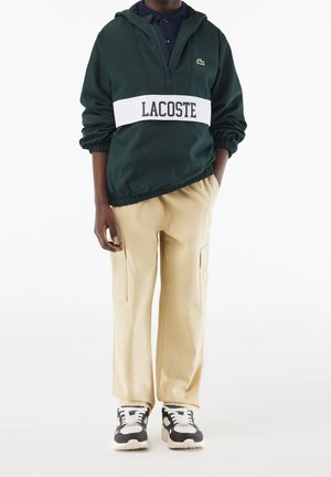 Person iført mørkegrøn Lacoste hættetrøje med hvidt logopanel, beige cargobukser og sorte og hvide sneakers, stående med den ene hånd i lommen.