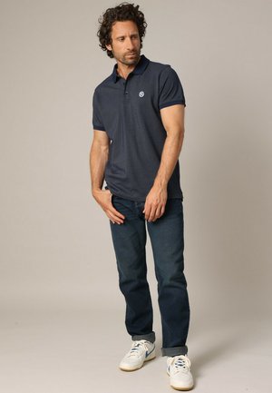 Polo shirt bleu marine avec un motif texturé et un col, associé à un jean bleu foncé et des baskets blanches avec des accents bleus.