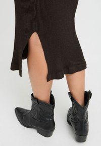Schwarze Leder-Cowboy-Stiefel mit spitzen Zehen, detaillierten Nähten und Ziehschlaufen, kombiniert mit einem braunen gerippten Kleid mit einem Seitenschlitz.