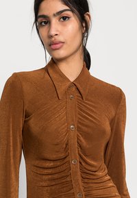 Camisa de dress de manga longa castanha, feita de um tecido suave com fecho de botão, apresentando detalhe franzido na frente e um colarinho pontiagudo.