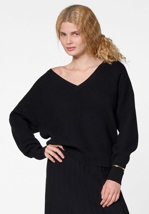Femme blonde portant un pull en V texturé noir à manches longues et une jupe noire côtelée, posant avec une main sur la hanche.