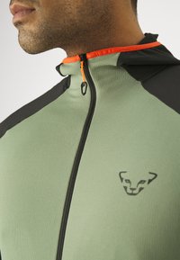Green zip-up jacket with black accents. Textured fabric; features an orange zipper and logo detail. Slim fit with a hooded collar.