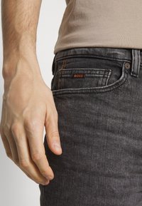 En närbild av en mans hand som vilar på midjan av grå jeans med en orange "BOSS" etikett synlig på fickan.