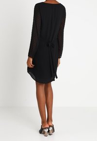 Robe noire avec de longues manches transparentes à pois, ceinture à la taille et ourlet fluide. Associée à des talons noirs avec des accents métalliques.