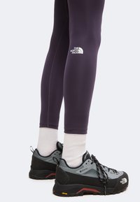 Άτομο που φοράει σκούρο κολάν North Face, λευκές κάλτσες και γκρι ορειβατικά παπούτσια North Face με μαύρες σόλες, στέκεται σε λευκό φόντο.