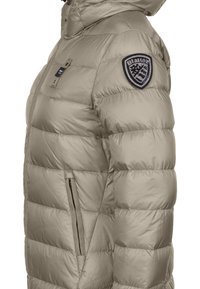 Vaalean beige puffer-takki, jossa on huppu, tikattu muotoilu, sivuvetoketjullinen tasku ja päällystetty logo vasemmassa hihassa. Sileä, kiiltävä pinta.