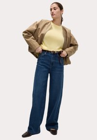 Veste bomber beige, pull jaune à col rond, jeans large taille haute bleu foncé, ceinture marron avec boucle, et chaussures foncées.