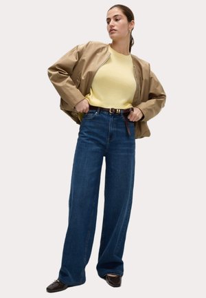Beige bomberjack, gele crewneck trui, high-waisted donkerblauwe wijde jeans, bruine riem met gesp en donkere schoenen.