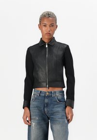 Persona con capelli ricci corti, che indossa una giacca nera con zip e maniche in pelle, insieme a jeans blu strappati, in piedi su uno sfondo bianco.