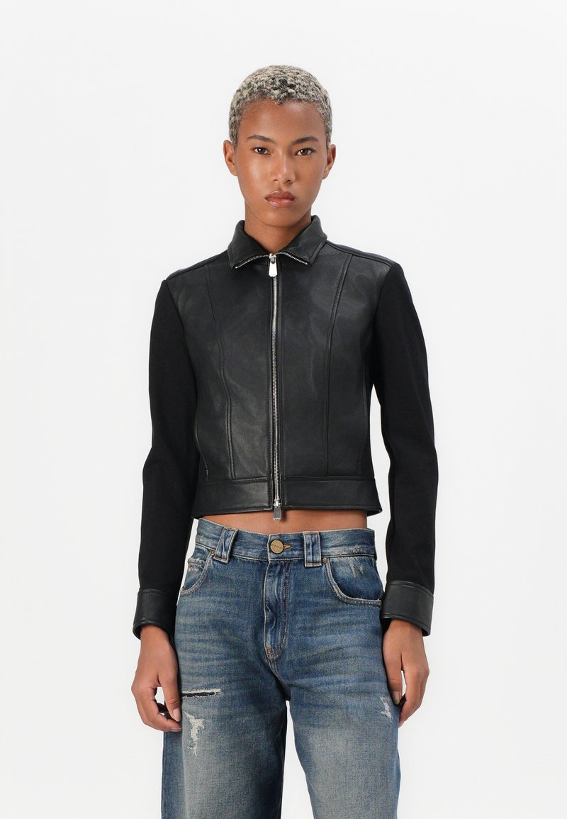 Persona con capelli ricci corti, che indossa una giacca nera con zip e maniche in pelle, insieme a jeans blu strappati, in piedi su uno sfondo bianco.