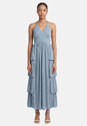 Donna che indossa un abito lungo azzurro chiaro senza maniche con scollo all’americana e dettagli a balze a strati, con décolleté aperte beige, in piedi su uno sfondo chiaro.