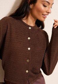 Brązowy sweter kardigan o fakturze, z długimi rękawami, okrągłym dekoltem i centralnym zapięciem na guziki w jasnym kolorze. Miękki materiał, swobodny krój.