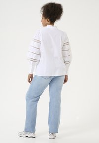 Blouse blanche avec des manches bouffantes volumineuses ornées de dentelle. Portée avec un jean en denim bleu clair et des baskets de teinte neutre.
