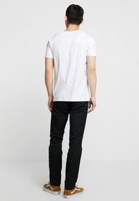 T-shirt bianca in cotone, maniche corte, design semplice, abbinata a pantaloni neri, che mostrano una vestibilità casual. Sneakers marroni e bianche completano l'outfit.