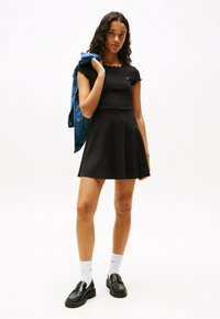 Schwarzes Kurzarmkleid mit tailliertem Oberteil, weitem Rock und Wellenkragen. Kombiniert mit einer Jeansjacke und schwarzen Slippern.