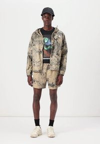 Leichte, beige Blümchenjacke mit Kapuze, schwarzes T-Shirt mit Grafik und passende Shorts. Sneakers mit dicke Sohlen, schwarze Socken.