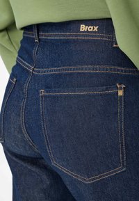 Dunkelblaue Denim-Jeans mit einer taillierten Passform, die orangefarbene Nähte, eine Gesäßtasche und ein Brax-Logo am Bund aufweisen.