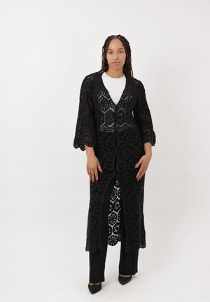 Frau mit langen Zöpfen trägt lange schwarze Häkel-Strickjacke über weißem Top und schwarzen Hosen, steht vor schlichtem Hintergrund.