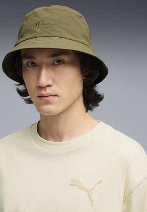 Joven con sombrero tipo bucket verde oliva de Puma y camiseta beige de Puma sobre un fondo de degradado gris.