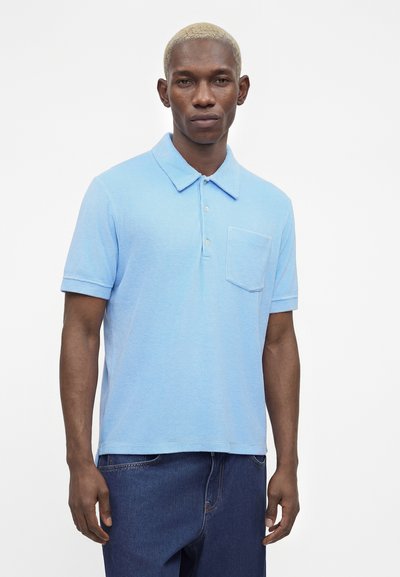 Άνδρας με κοντά ξανθά μαλλιά που φοράει ανοιχτό μπλε κοντομάνικη πόλο μπλούζα με τσέπη στο στήθος και σκούρο μπλε τζιν, στέκεται μπροστά σε λευκό φόντο.