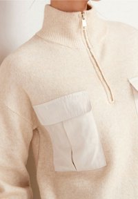 Polo beige à fermeture éclair avec un col haut côtelé. Comprend deux grandes poches plates en tissu beige plus clair. Matière douce et texturée.