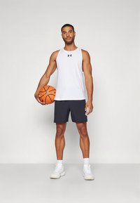 Weißes ärmelloses Sporttop mit schwarzem Logo, kombiniert mit dunklen Shorts und weißen Turnschuhen; hält einen Basketball. Einfaches und funktionales Design.