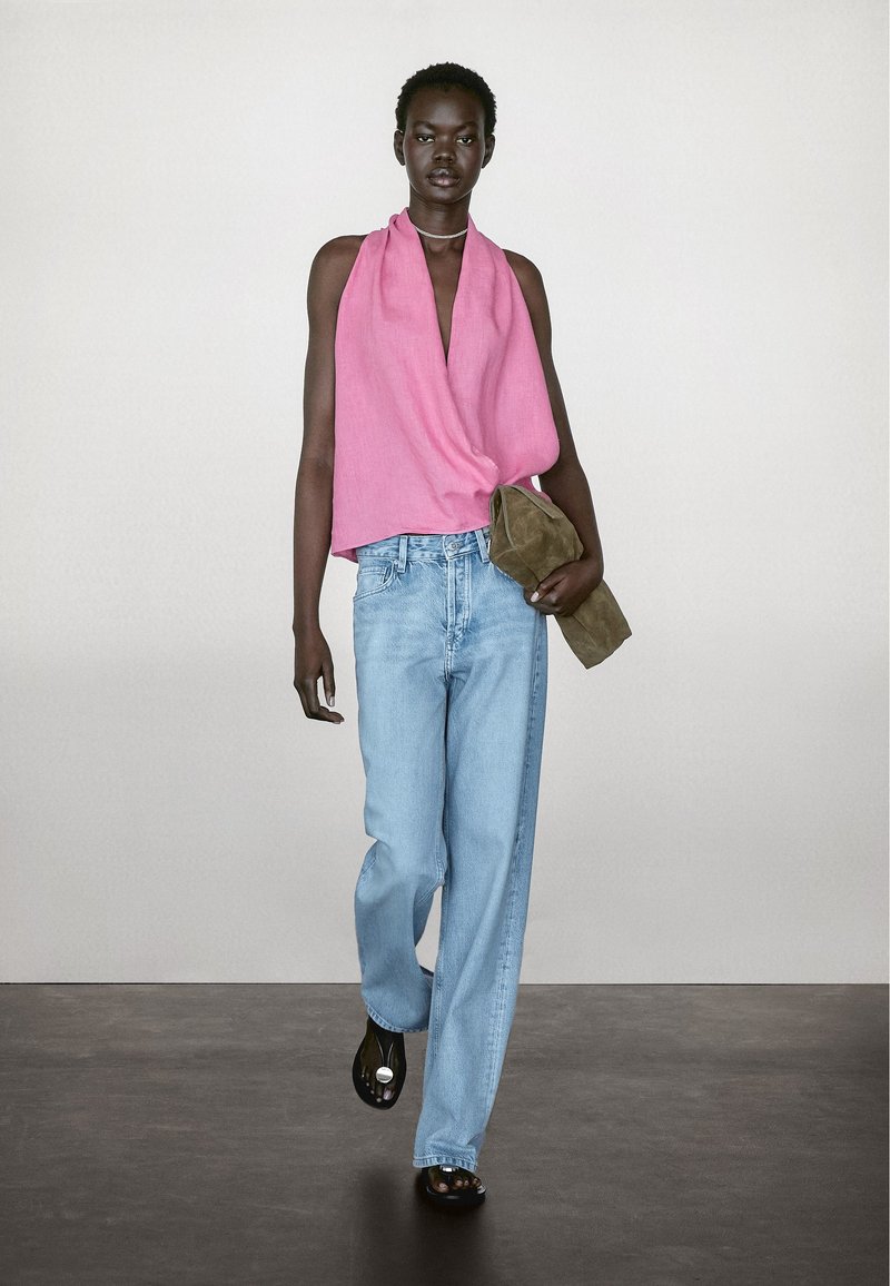 Różowy top bez rękawów o drapowanej konstrukcji, jasnoniebieskie dżinsy proste, i zielona torebka z zamszu, noszone z czarnymi butami. Proste tło.