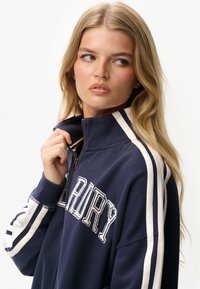 Sudadera navy con cremallera y acentos de rayas blancas, con letras en la parte frontal. Cuenta con un cuello alto y puños de punto, fabricada en un tejido suave.