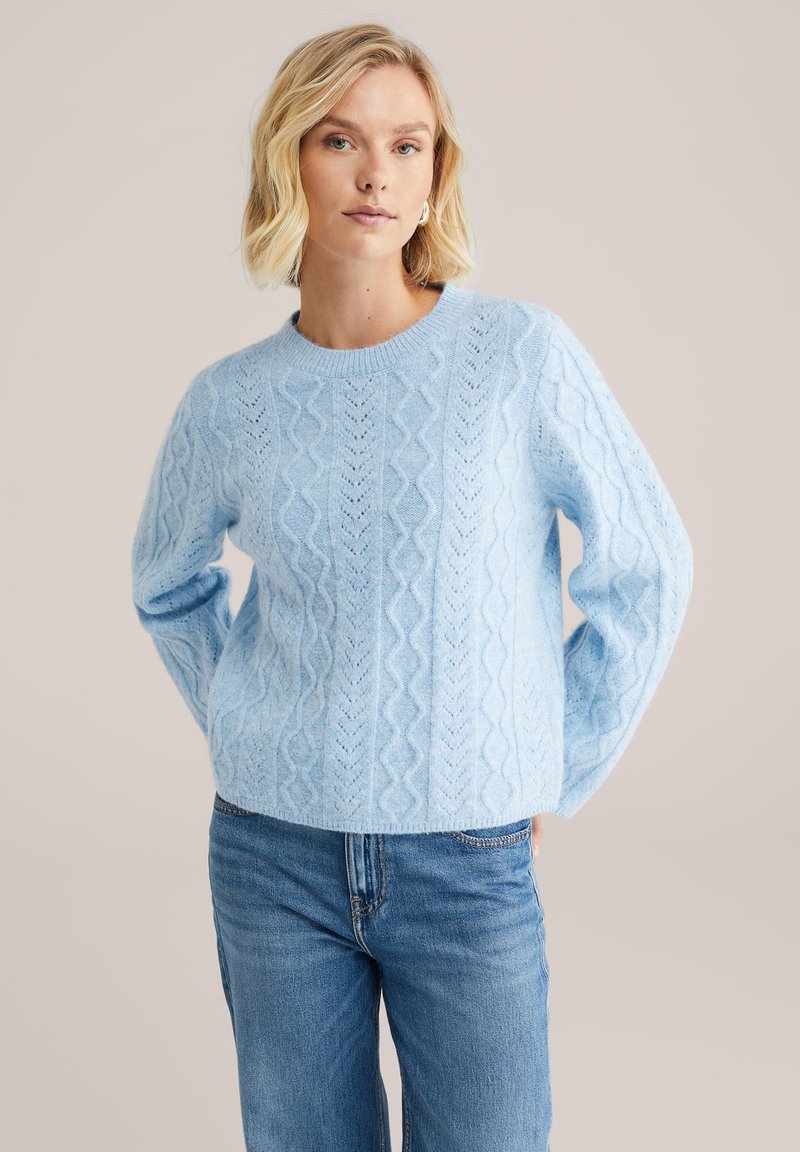 Pulover din tricotare cu fir albastru deschis, cu guler rotund și mâneci lungi. Prezintă modele texturate și o croială lejeră. Se purta cu blugi albaștri.