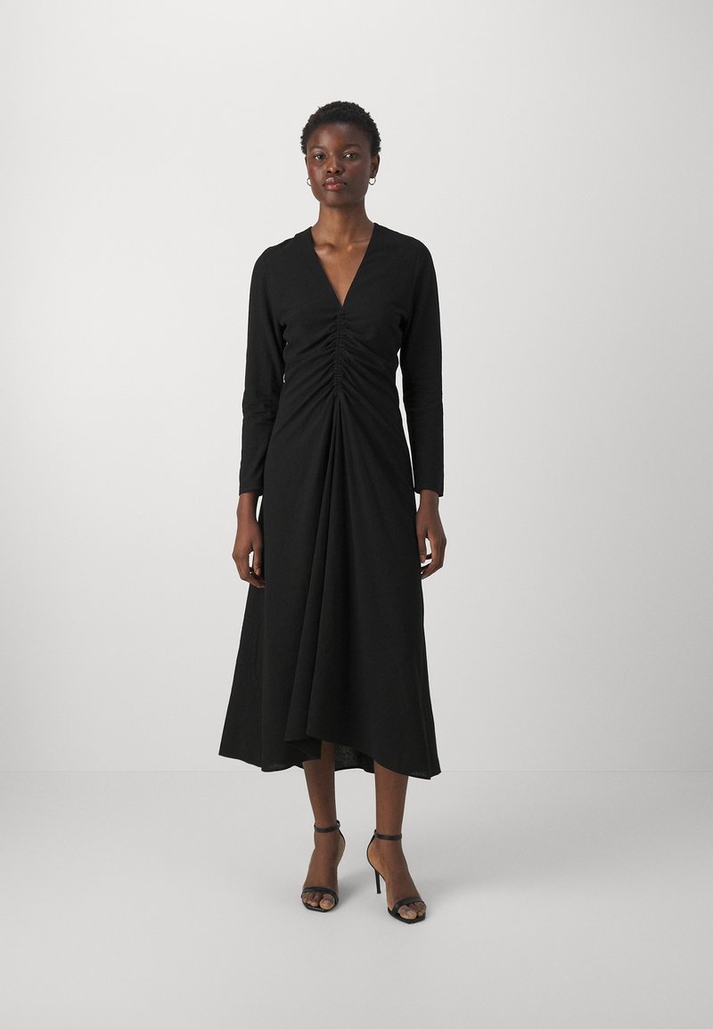 Vestido negro con mangas largas y un escote en V profundo. Detalle fruncido en la parte frontal que realza la silueta, falda de longitud media fluida, llevado con sandalias de tacón negras.