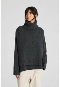 Naine, kes kannab tumedat halli üle suurusega kaelusega kampsunit ja beeže pükse, seisab ühevärvilise heleda tausta ees, vaadates küljele.